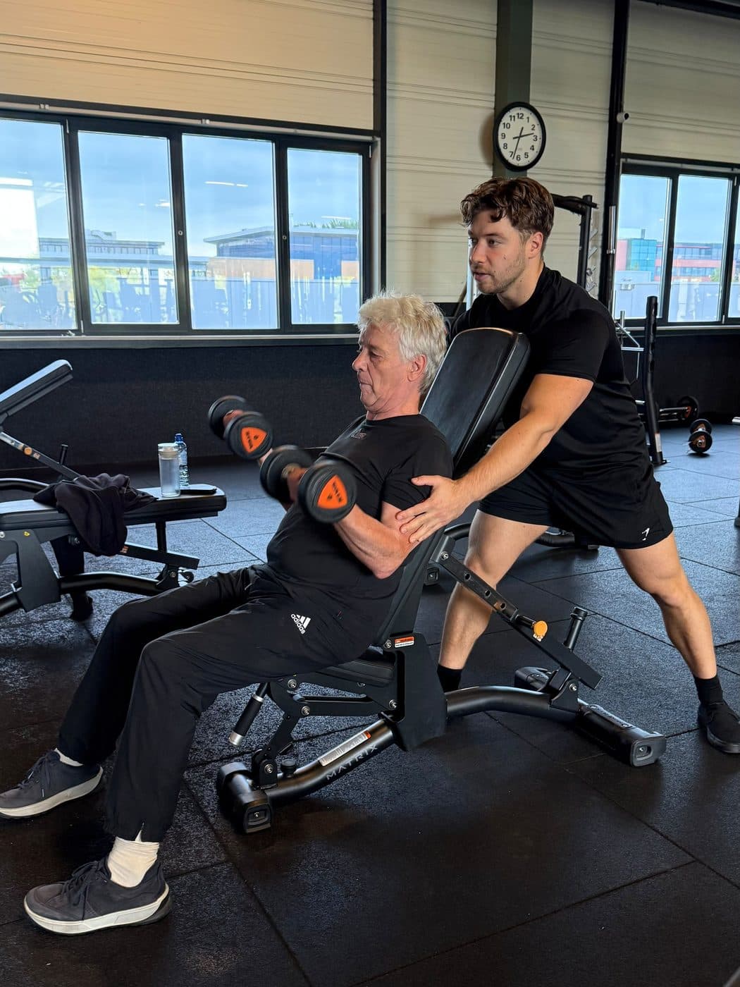 Rian staat achter een klant van in de zestig en corrigeert zijn dumbbellpress